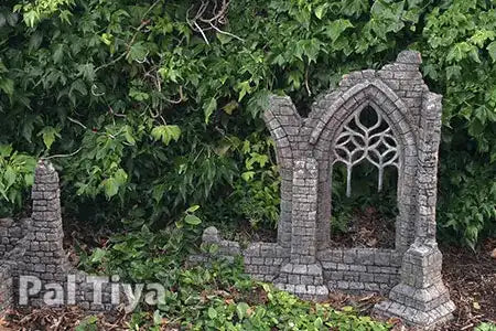 A miniature gray stone gothic ruins garden stake with an arched window design, set against a background of green garden foliage. A 'Pal Tiya' brand watermark is visible in the bottom left corner.