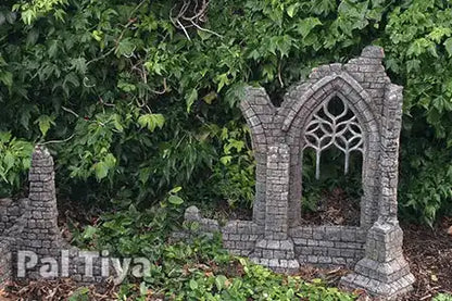 A miniature gray stone gothic ruins garden stake with an arched window design, set against a background of green garden foliage. A 'Pal Tiya' brand watermark is visible in the bottom left corner.