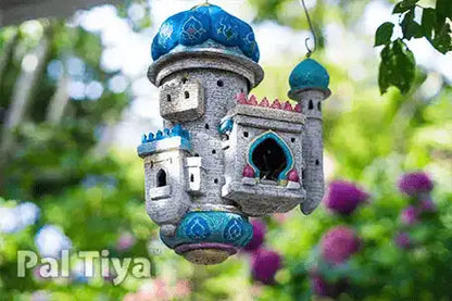 A decorative hanging birdhouse shaped like a miniature castle, with blue domes, a gray stone-textured body, and pink accents, photographed outdoors against a blurred green garden background. The brand name 'Pal Tiya' appears in the lower left corner.