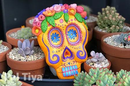 A colorful decorative skull ornament with floral details on top, displayed among small potted succulents and cacti. Made with Pal Tiya Premium.