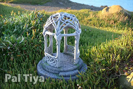 A miniature white fairy garden gazebo with intricate leafy vine detailing on a stone-textured base, set in a grassy outdoor environment. A watermark reading 'Pal Tiya' is visible in the bottom left corner.