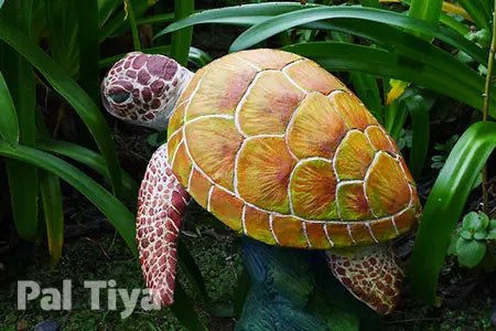 A decorative sea turtle garden statue with a yellow-orange patterned shell and pinkish-brown patterned flippers, displayed among green artificial foliage. The brand name 'Pal Tiya' is visible in the bottom-left corner.
