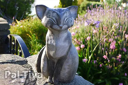 A gray cat garden statue with closed eyes sits on a stone ledge in a sunlit garden with green foliage and pink flowers in the background. Made with Pal Tiya Premium