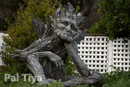 A weathered tree-troll garden statue with a calm face and branches for limbs, placed outdoors among green shrubs in front of a white lattice fence. A 'Pal Tiya' watermark is visible in the lower left corner.